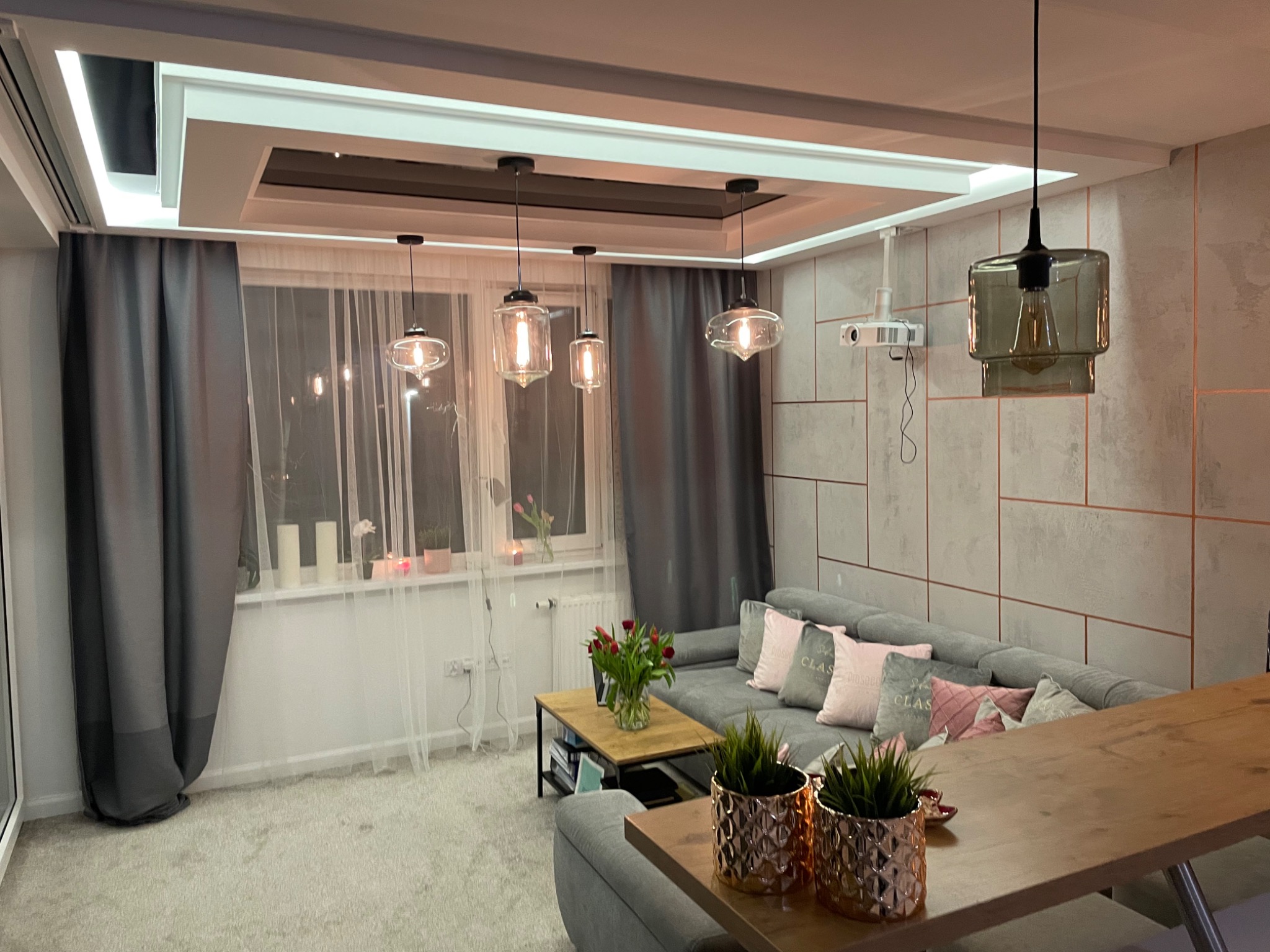 Nowoczesny salon z szarą sofą, drewnianym stolikiem kawowym i dekoracyjnymi lampami wiszącymi, z projektorem zamontowanym na ścianie.