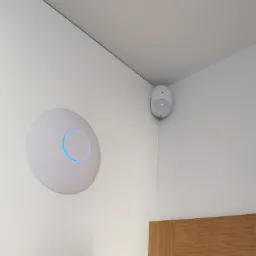 Biały punkt dostępowy Wi-Fi z niebieskim podświetleniem obok czujnika ruchu zamontowanego w rogu pomieszczenia, widoczne fragmenty ościeżnicy drzwi.