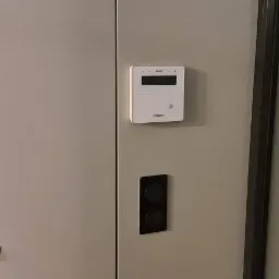 Biały panel sterowania systemem alarmowym marki Satel, zamontowany na szarej ścianie obok czarnego podwójnego włącznika światła, z widocznymi drzwiami szafy z jasnobrązowymi skórzanymi uchwytami.