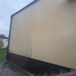 Fragment elewacji budynku z widocznym efektem mycia: część ściany czysta, część zabrudzona, ukazująca różnicę przed i po czyszczeniu. Widoczne schody i fragment trawnika.