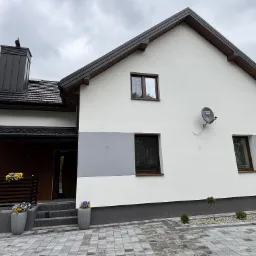 Dwukondygnacyjny dom z białą elewacją i szarym pasem akcentującym, ciemna dachówka, komin i stolarka okienna. Uwagę zwracają donice z kwiatami przy wejściu.