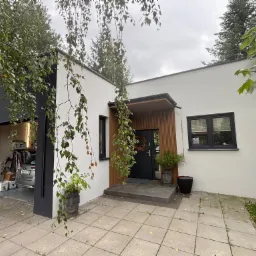 Nowoczesny dom z białą elewacją i czarnymi akcentami, widok z zewnątrz, z garażem i roślinnością. Wieniec na drzwiach wejściowych i donice w kształcie głowy.