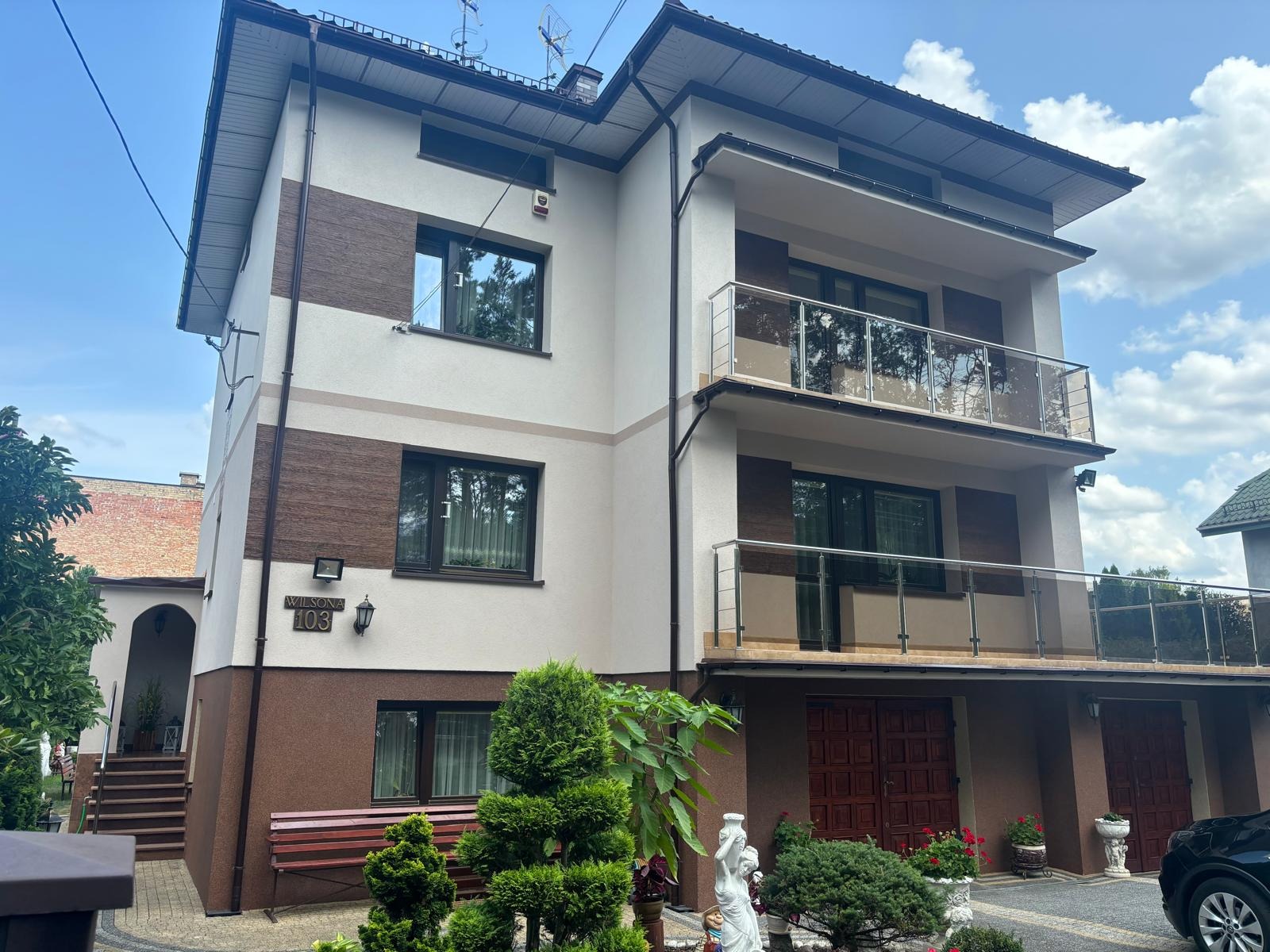 Dwupiętrowy dom z beżowo-brązową elewacją, balkonami z przeszklonymi balustradami, numerem domu 'WILSONA 103' i zadbanym ogrodem z rzeźbą.