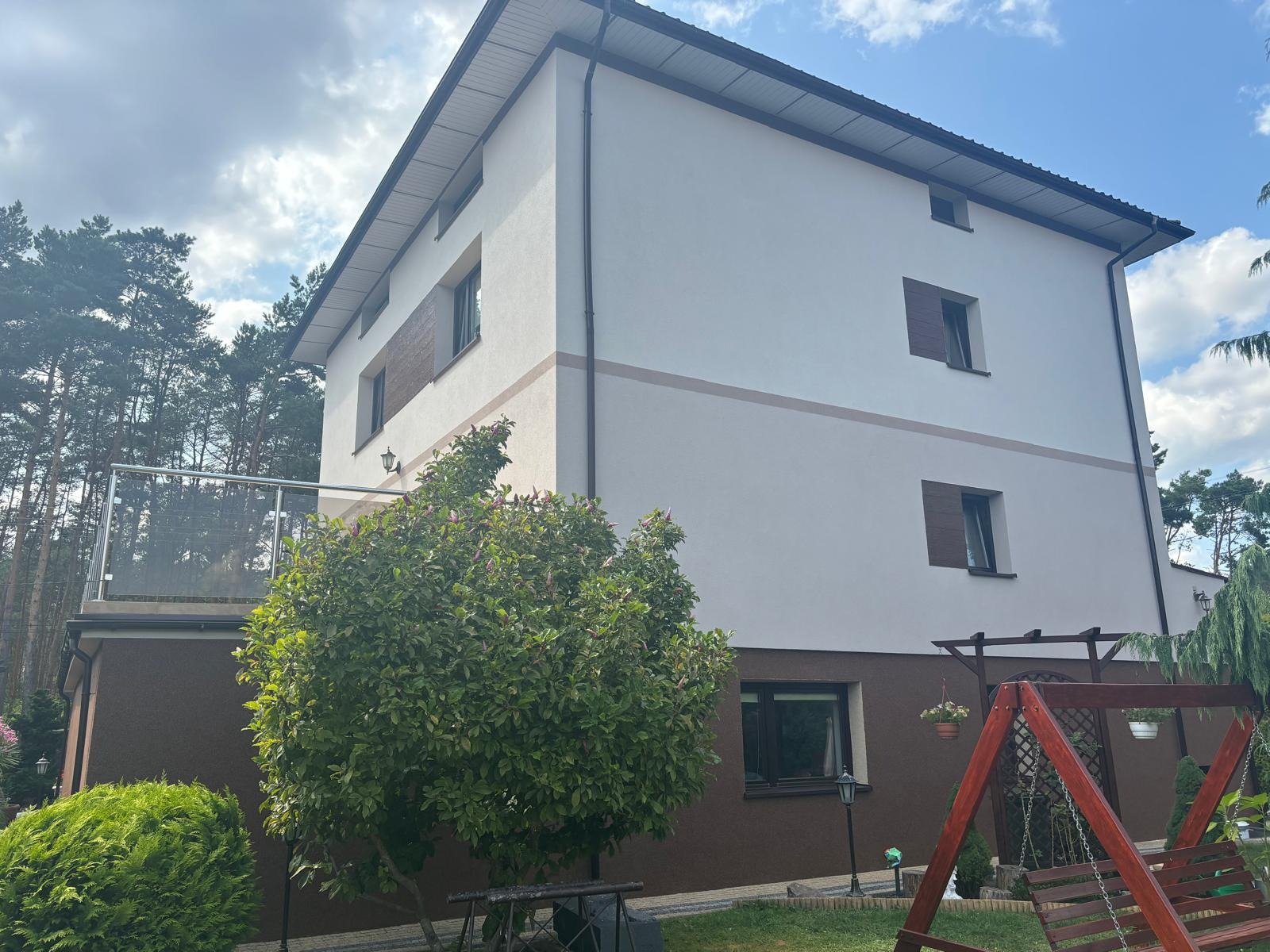 Dwukondygnacyjny dom z białą elewacją, brązowym pasem i ciemnobrązową dolną częścią, widoczny z ogrodu z drzewami i huśtawką