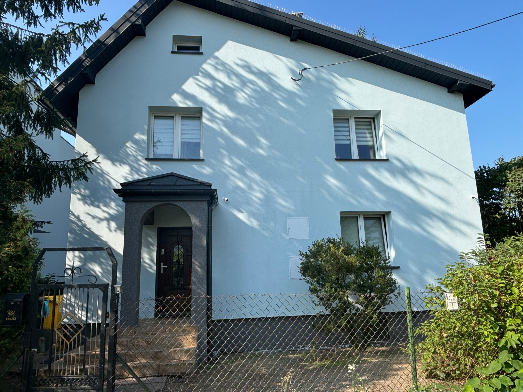 Jasno-niebieski dom jednorodzinny z ciemnym dachem i zabezpieczeniami przeciw ptakom, widziany zza ogrodzenia z siatki, z wyraźnymi cieniami drzew na elewacji.