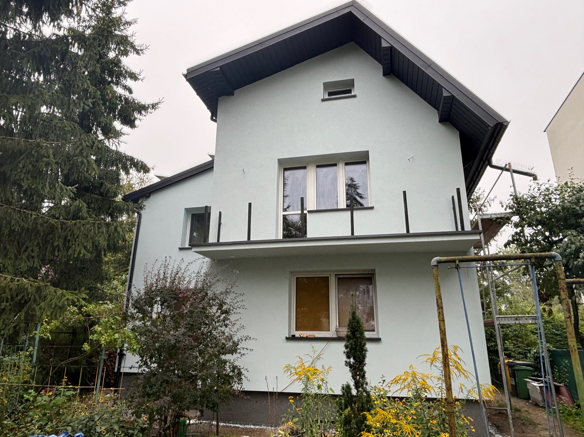 Dwupiętrowy dom z jasną elewacją i ciemnym dachem, widoczny z boku ogrodu z żółtymi kwiatami i metalową konstrukcją rusztowania.