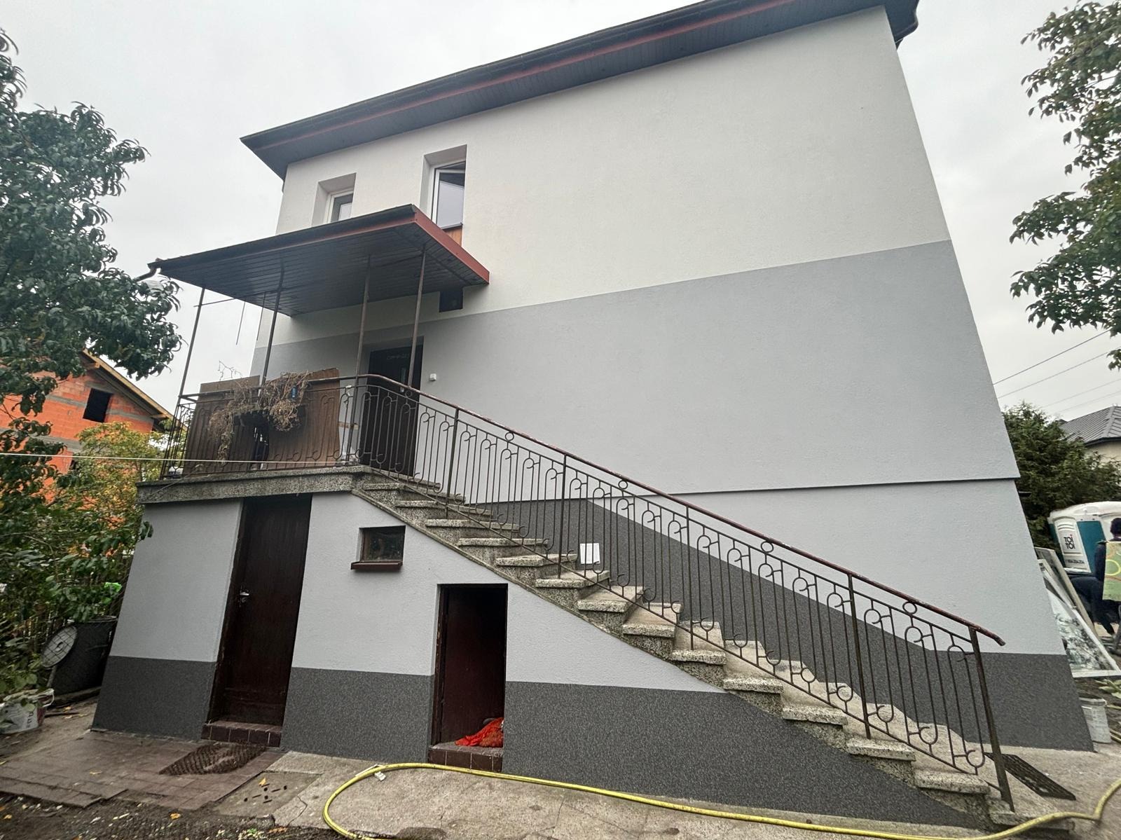 Dwukolorowa elewacja domu z zewnętrznymi schodami, widoczne świeże tynki i częściowo pomalowane fragmenty, żółty wąż ogrodowy na ziemi.