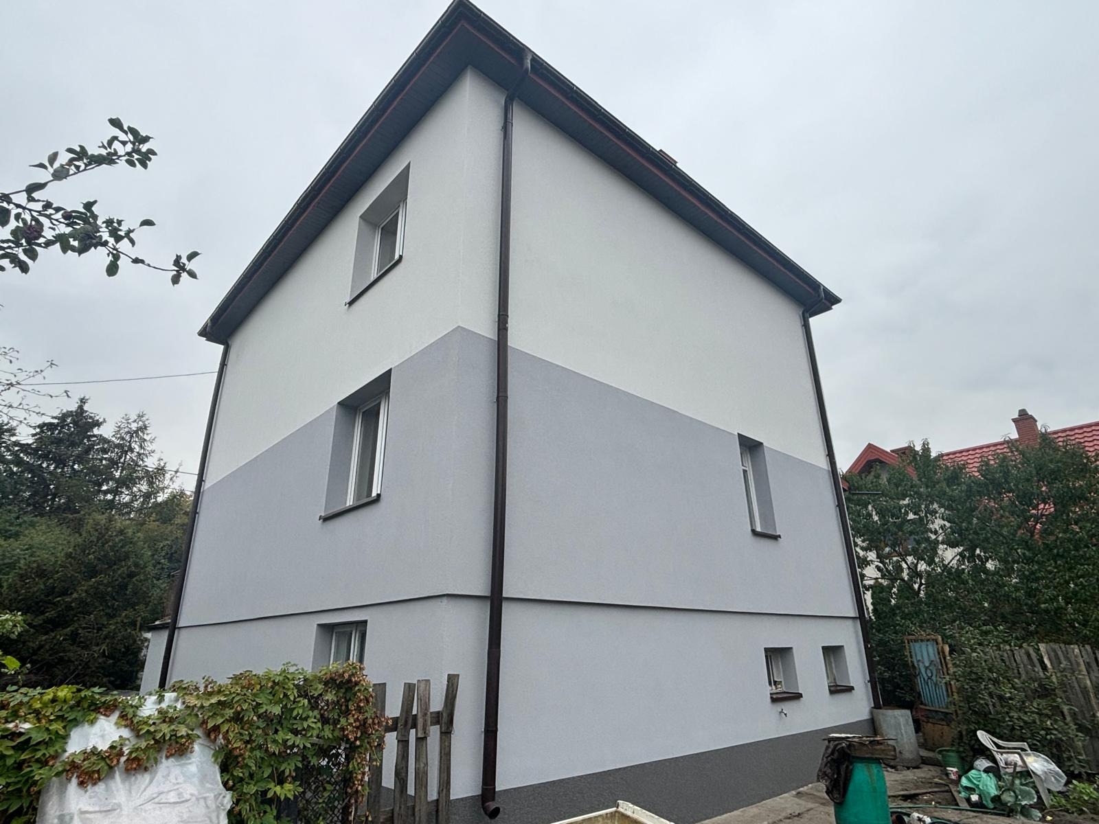 Dwukondygnacyjny dom z elewacją w dwóch odcieniach szarości, widok z dołu w pochmurny dzień, z widocznym fragmentem drewnianego płotu i roślinności.