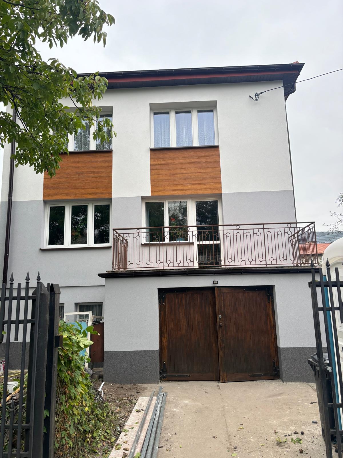 Dwukondygnacyjny dom z szarą i białą elewacją, z drewnianymi panelami dekoracyjnymi, balkonem z metalową balustradą i drewnianymi drzwiami garażowymi, widok z zewnątrz.