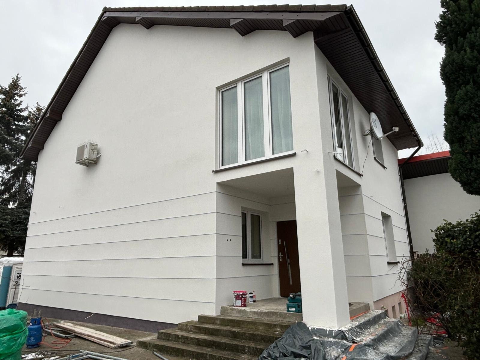 Dwupiętrowy dom z jasną elewacją z poziomymi pasami, widoczny klimatyzator, antena satelitarna, puszki z farbą i narzędzia na schodach.