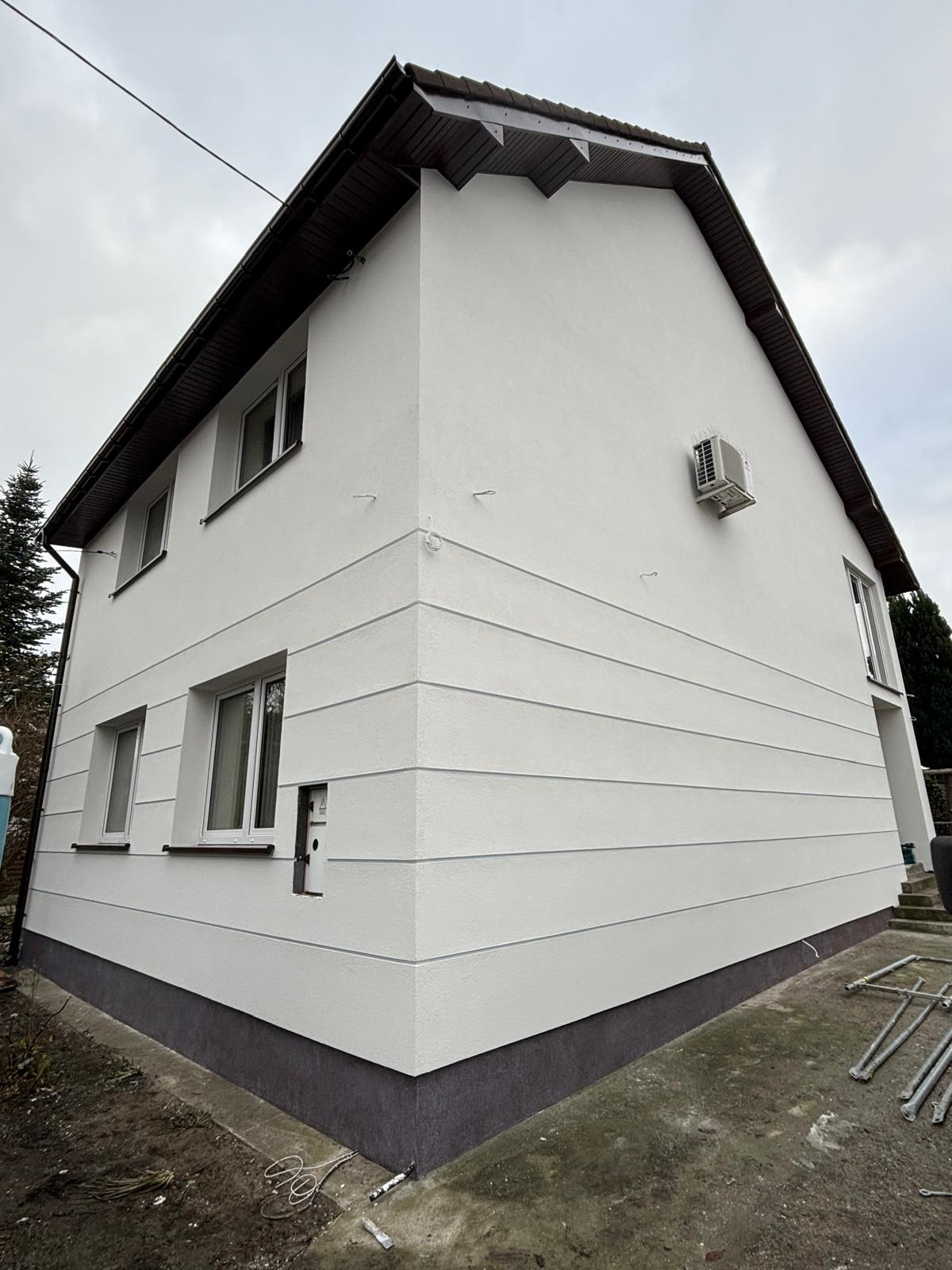 Dwupiętrowy dom po remoncie elewacji, z białymi ścianami z poziomymi pasami i ciemną podmurówką, widoczny klimatyzator na ścianie, z niedokończonym otworem na licznik i elementami rusztowania...