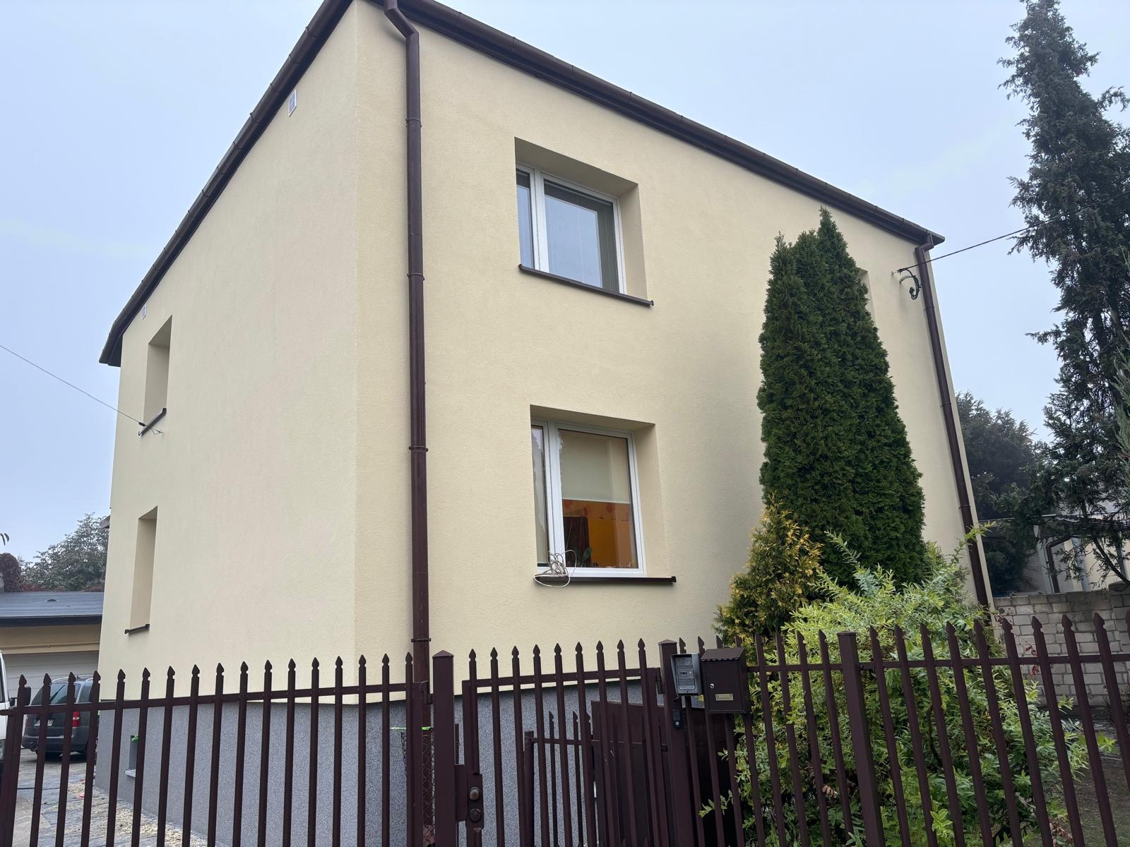 Dwupiętrowy dom z beżową elewacją i brązowymi rynnami, widoczny zza brązowego ogrodzenia z metalowych prętów, z tujami obok budynku.