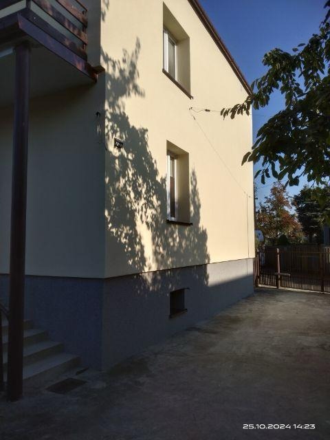 Dwupiętrowy dom z beżową elewacją i ciemnoszarą podmurówką, widziany z boku, z cieniem drzewa na ścianie.