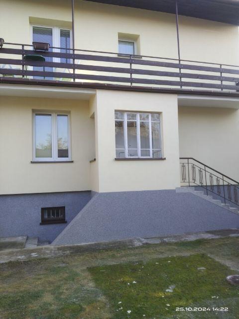 Dwukondygnacyjny dom z żółtą elewacją i szarą podmurówką z widoczną fakturą, balkon z drewnianą balustradą, schody z metalową barierką prowadzące do wejścia.