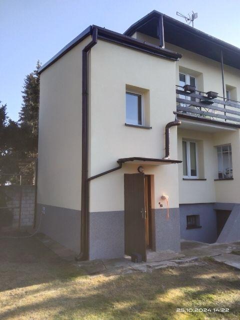 Dwukondygnacyjny budynek z elewacją w odcieniach beżu i szarości, widoczne rynny w kolorze brązowym, otwarte drzwi wejściowe i trawnik w słoneczny dzień.