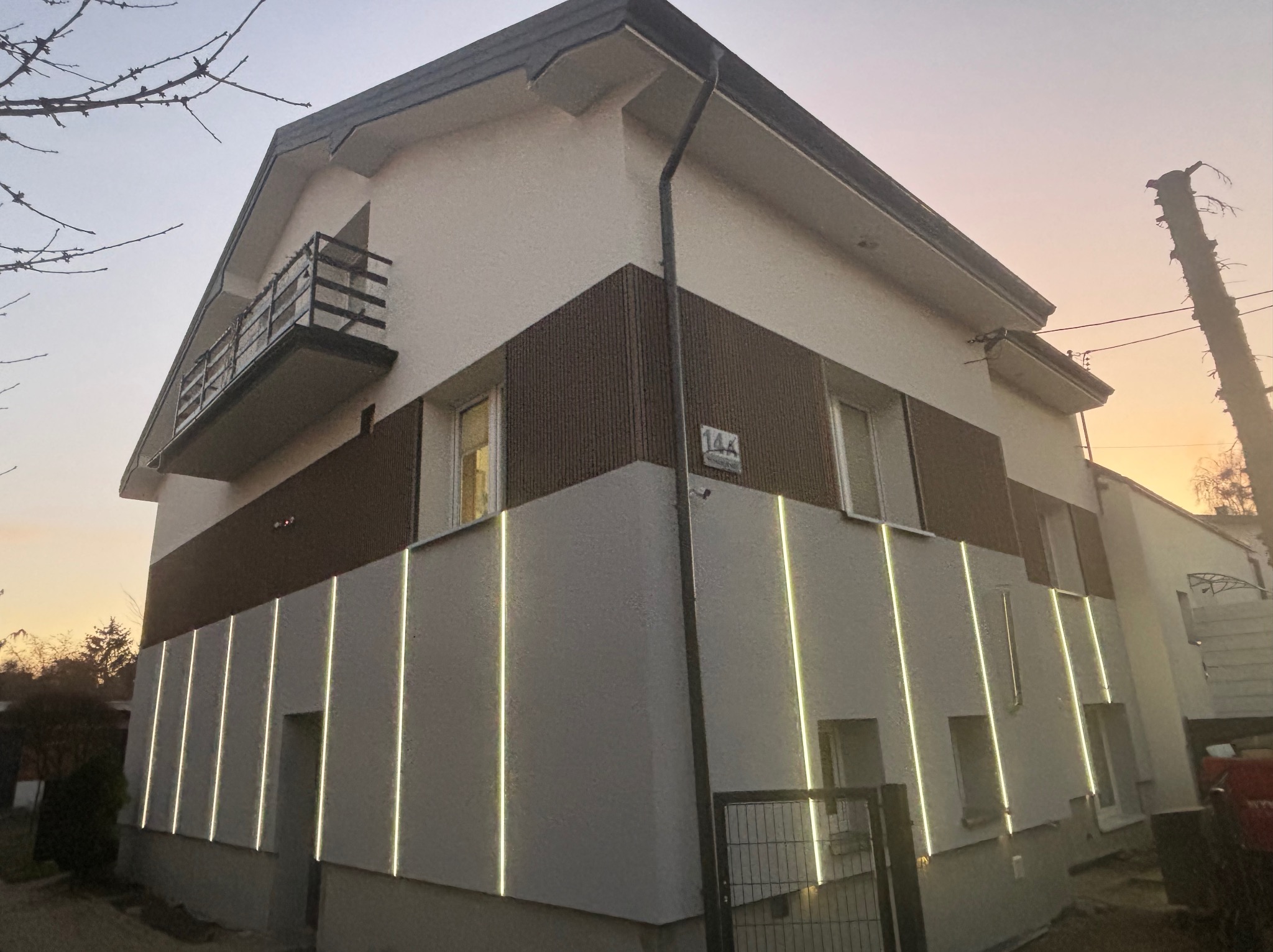 Dwukondygnacyjny dom z jasną elewacją, brązowym pasem na wysokości okien i pionowymi paskami świetlnymi, oświetlony ciepłym światłem zachodzącego słońca.