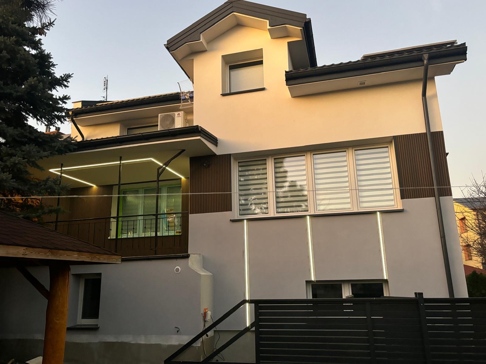 Dwukondygnacyjny dom z nową elewacją w odcieniach szarości i beżu, z oświetleniem LED podkreślającym pionowe i poziome linie, widok z zewnątrz na ogród.