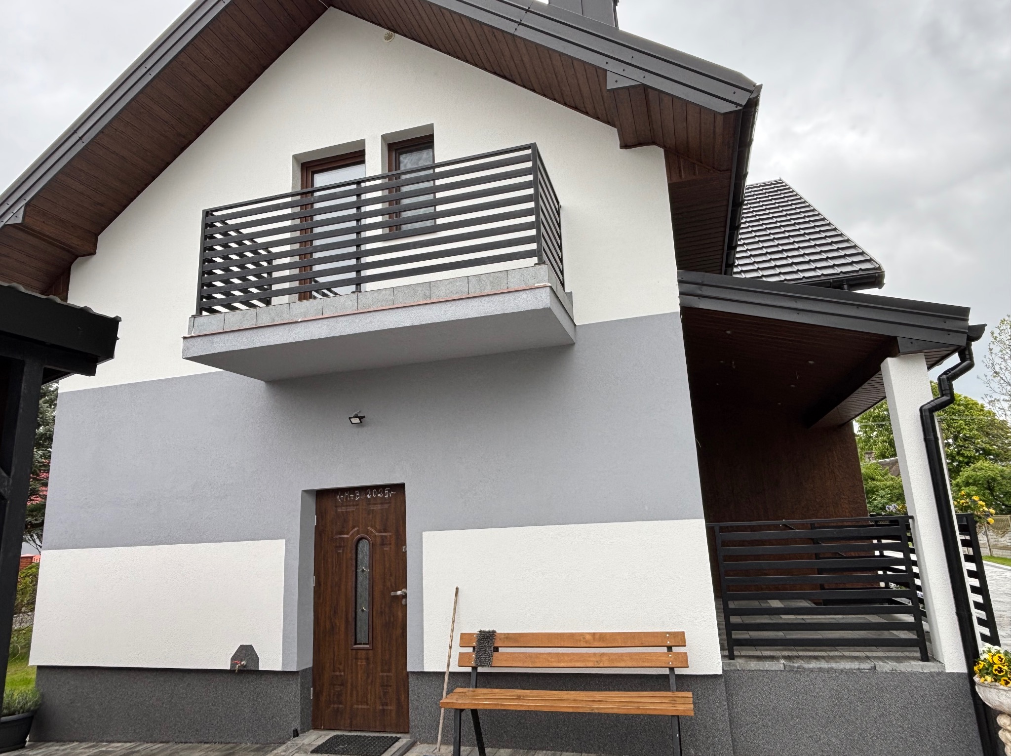 Dwukondygnacyjny dom z balkonem, elewacja w odcieniach bieli i szarości. Widoczny fragment dachu z brązową blachodachówką i drewniana ławka przed wejściem.