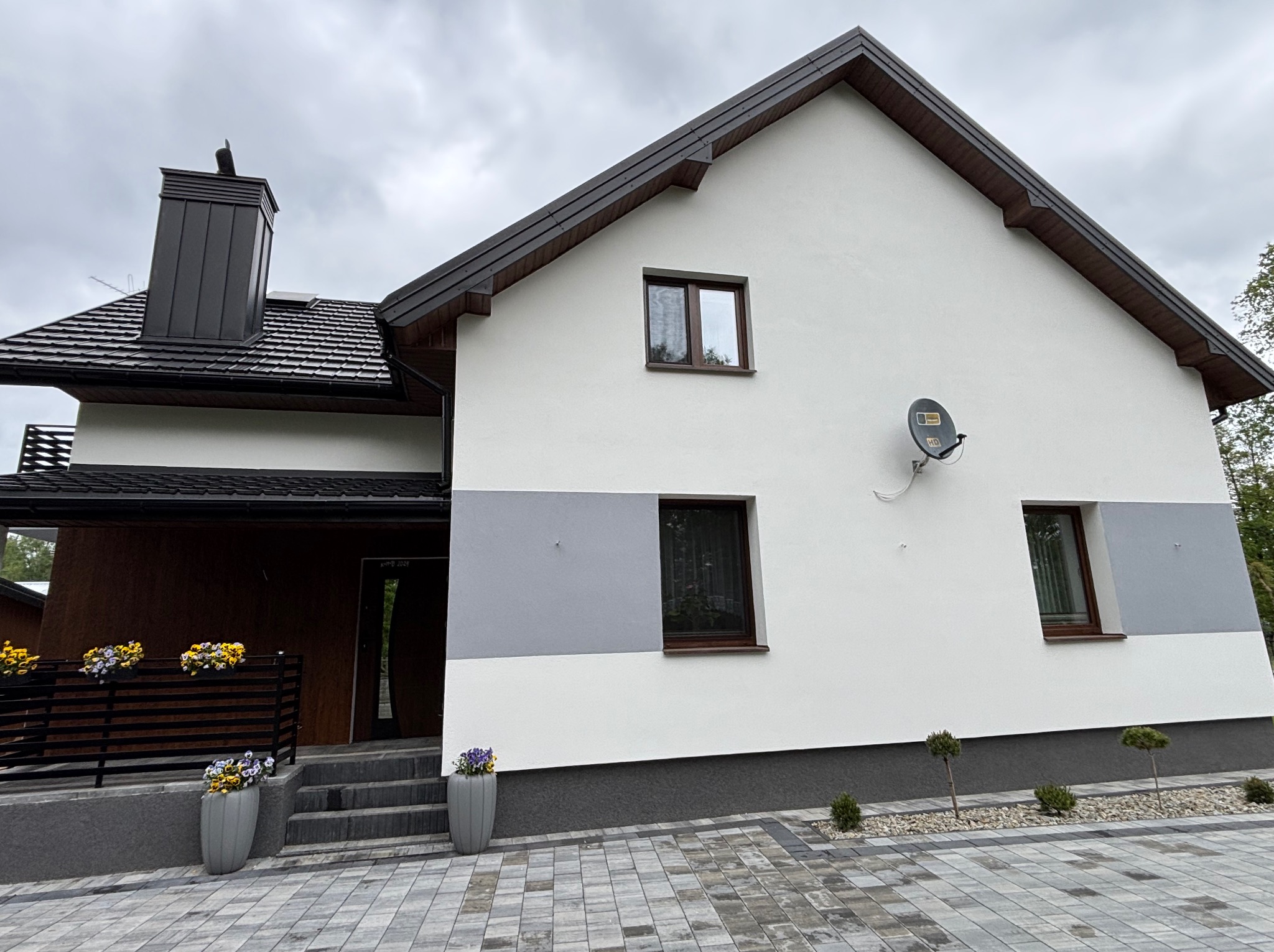 Dwukondygnacyjny dom z białą elewacją i szarym pasem akcentującym, ciemna dachówka, komin i stolarka okienna. Uwagę zwracają donice z kwiatami przy wejściu.