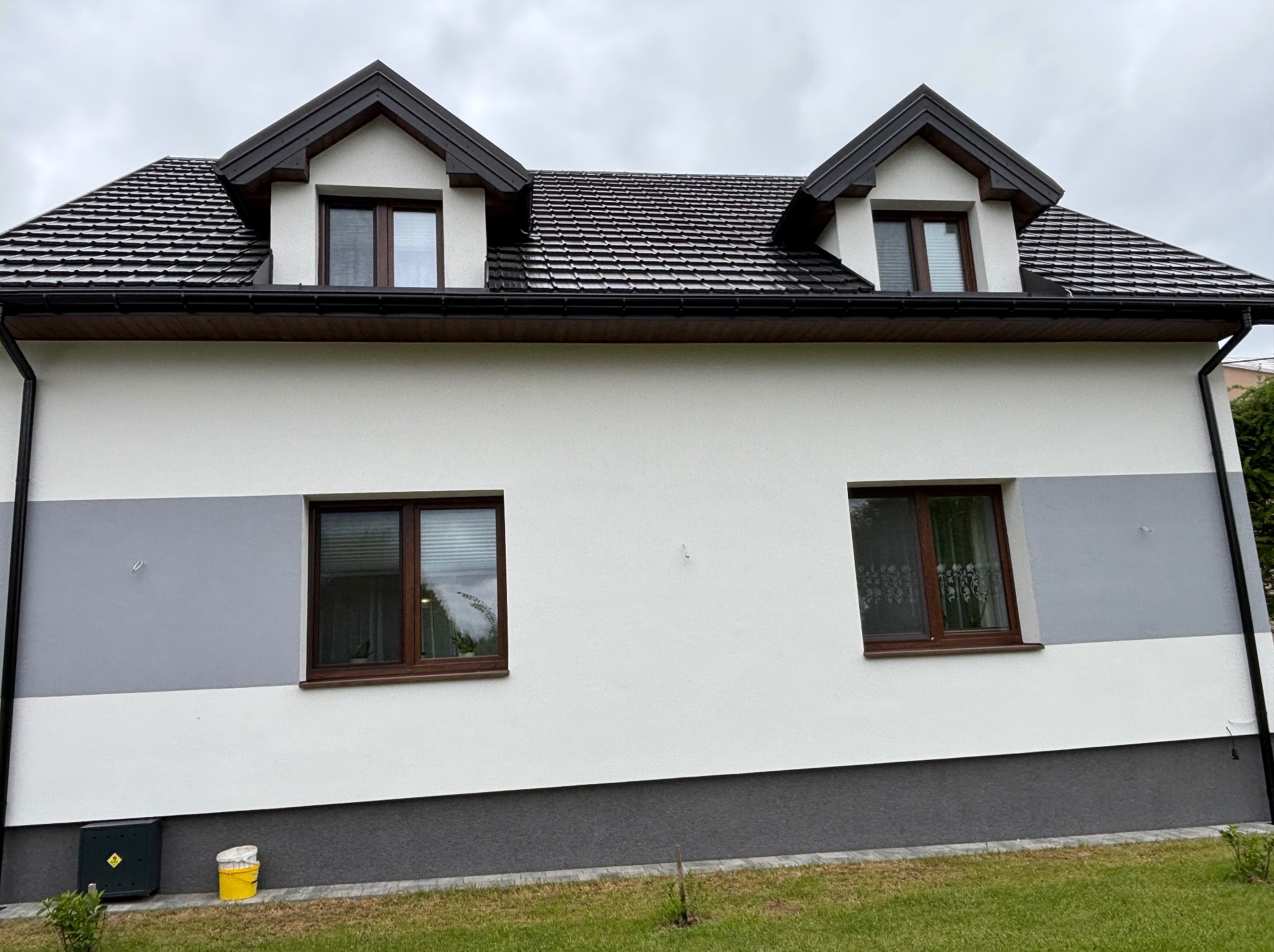 Dwukondygnacyjny dom z białą elewacją, szarymi pasami i brązowymi oknami. Widoczne dwa okna dachowe i fragment zielonego trawnika z przodu.