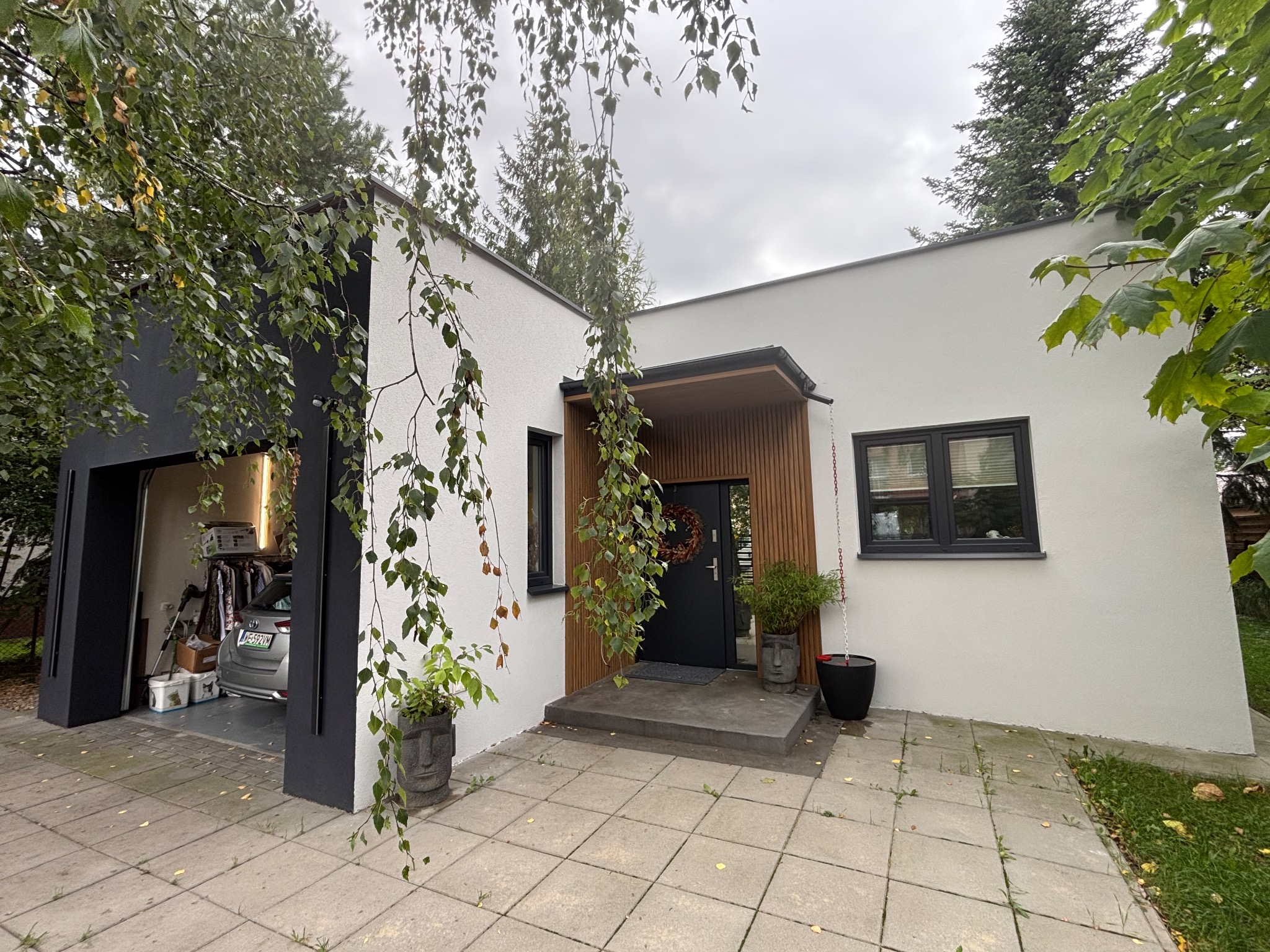 Nowoczesny dom z białą elewacją i czarnymi akcentami, widok z zewnątrz, z garażem i roślinnością. Wieniec na drzwiach wejściowych i donice w kształcie głowy.