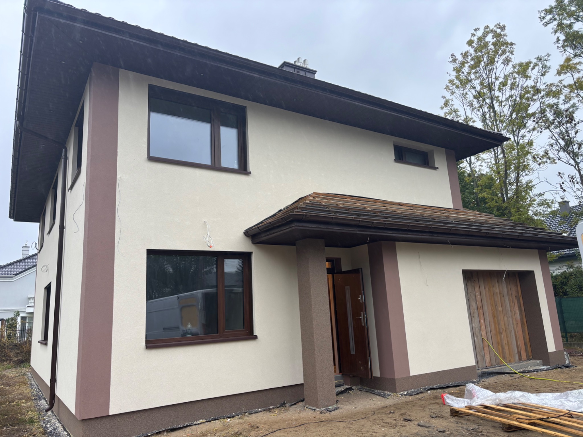 Dwukondygnacyjny dom z jasną elewacją i brązowymi akcentami, widoczny z boku, z otwartymi drzwiami wejściowymi i elementami drewnianymi.