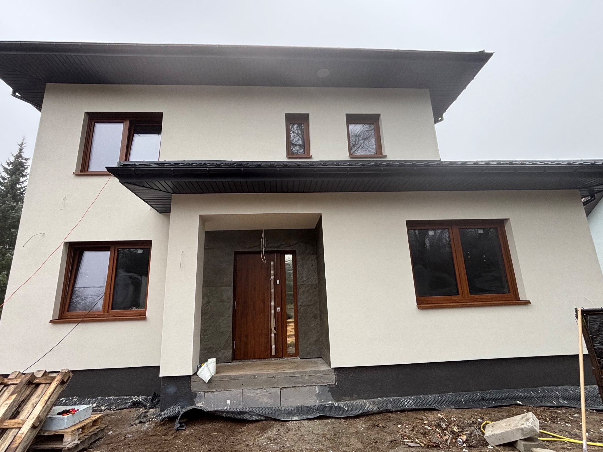 Dwukondygnacyjny dom z jasną elewacją, brązowymi oknami i drzwiami. Widoczne detale wykończenia, czarny dach i elementy otoczenia w trakcie prac.