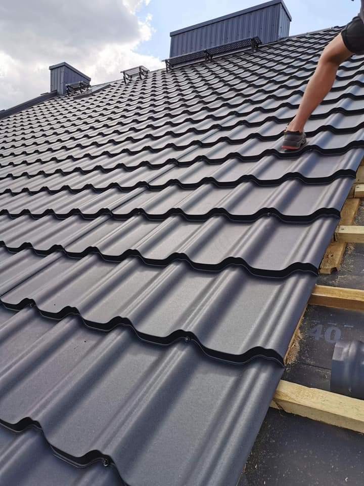 Montaż blachodachówki w kolorze grafitowym na drewnianej konstrukcji, widoczna noga dekarza w butach roboczych, fragment dachu z kominem w tle, pochmurne niebo.
