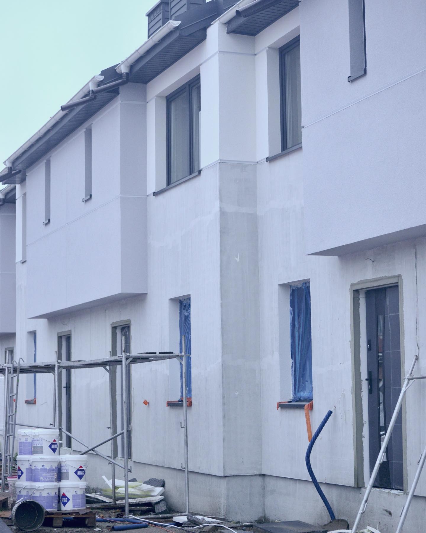 Nowe domy w zabudowie szeregowej w trakcie wykańczania elewacji, widoczne rusztowania, farby Atlas, okna zabezpieczone folią, wystające rurki instalacyjne.