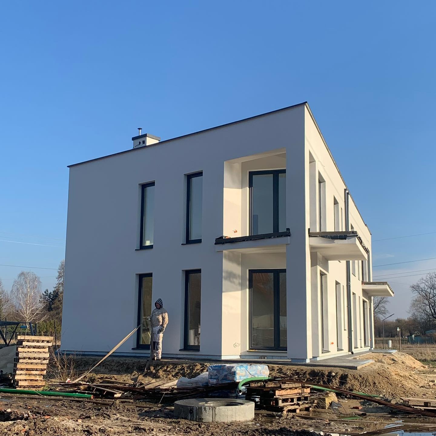 Nowoczesny, dwupiętrowy dom z płaskim dachem, świeżo otynkowany na biało, z ciemnymi ramami okiennymi, w trakcie prac wykończeniowych na zewnątrz, widoczny pracownik w kombinezonie ochronnym...