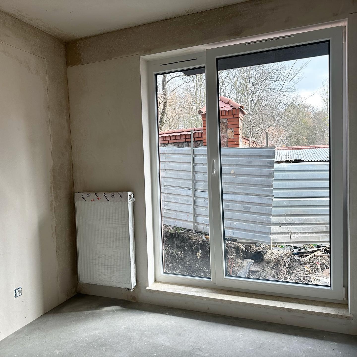 Wnętrze w stanie deweloperskim z zamontowanym, ale jeszcze ofoliowanym grzejnikiem i dużym oknem przesuwnym z widokiem na podwórze z metalowym ogrodzeniem.
