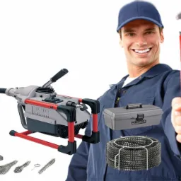 Uśmiechnięty hydraulik w niebieskim uniformie prezentuje zestaw narzędzi do udrażniania rur: maszynę Ridgid Kollmann, sprężynę w metalowym koszu, wymienne końcówki i skrzynkę narzędziową, trzymając...