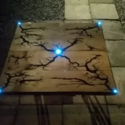 Drewniana płyta z wypalonym wzorem przypominającym drzewo, podświetlona niebieskimi diodami w rogach i w centrum, leżąca na kamiennej posadzce.