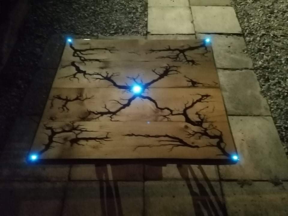 Drewniana płyta z wypalonym wzorem przypominającym drzewo, podświetlona niebieskimi diodami w rogach i w centrum, leżąca na kamiennej posadzce.