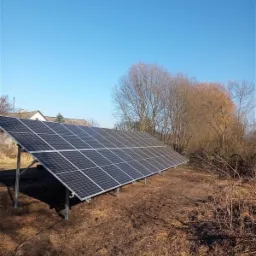 Panel słoneczny na metalowej konstrukcji, zainstalowany na nierównym, brązowym gruncie, z widocznym fragmentem dachu budynku w tle i błękitnym niebem.