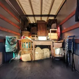 Wnętrze samochodu dostawczego wypełnione meblami i przedmiotami, w tym krzesłami, stołem, szafą i wiklinowymi koszami, zabezpieczonymi na czas transportu.