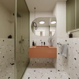 Nowoczesna łazienka z kabiną prysznicową oddzieloną szkłem ryflowanym, umywalką w kolorze terakoty i lustrem o zaokrąglonym kształcie. Na ścianach i podłodze płytki imitujące lastryko.