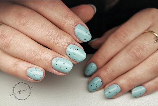 Dłonie z pomalowanymi na jasnoniebiesko paznokciami z czarnymi kropkami, imitującymi ptasie jajko, na serdecznym palcu złoty pierścionek.