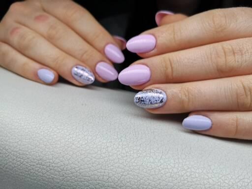 Dłonie z owalnymi paznokciami w odcieniach fioletu i lila, jeden paznokieć z srebrnym zdobieniem, na jasnym tle.