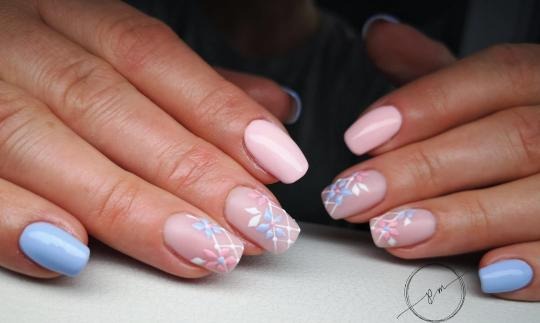 Dłonie z pastelowymi paznokciami; jeden paznokieć niebieski, pozostałe w odcieniach różu z ręcznie malowanymi kwiatowymi wzorami w błękicie i różu na siatce z białych linii.