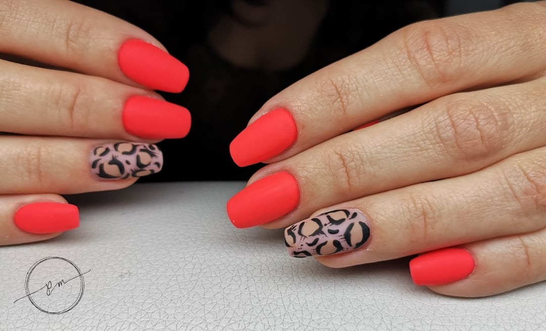 Zbliżenie dłoni z pomalowanymi na matowy koralowy kolor paznokciami, z akcentem panterki na paznokciach serdecznych, ułożonych na teksturowanym białym tle.