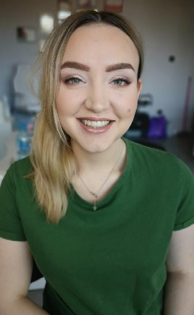 Uśmiechnięta blondynka z delikatnym makijażem dziennym, podkreślone brwi i błyszczące usta, na szyi srebrny naszyjnik z małą zawieszką, ubrana w zielony t-shirt.