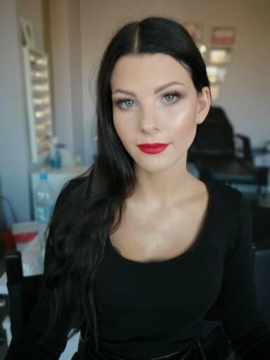 Portret kobiety z wyrazistym makijażem: czerwone usta, podkreślone oczy, rozświetlona cera i kruczoczarne włosy.