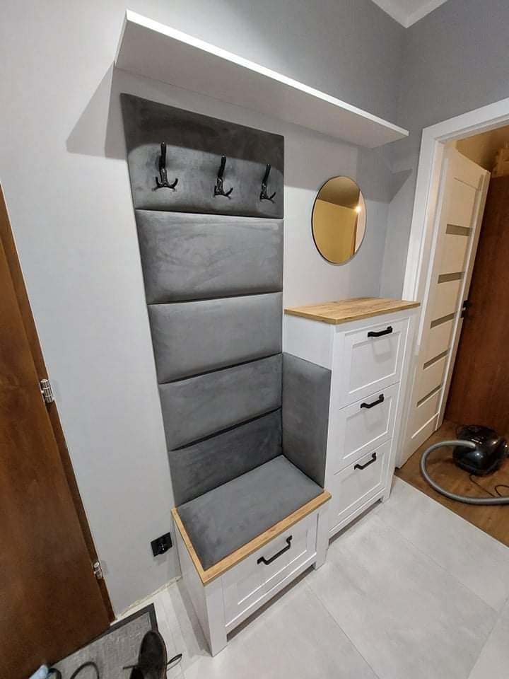 Nowoczesny przedpokój z tapicerowanym panelem ściennym z wieszakami, szafką na buty z drewnianym blatem i okrągłym lustrem, uzupełniony o białą półkę i odkurzacz w tle.