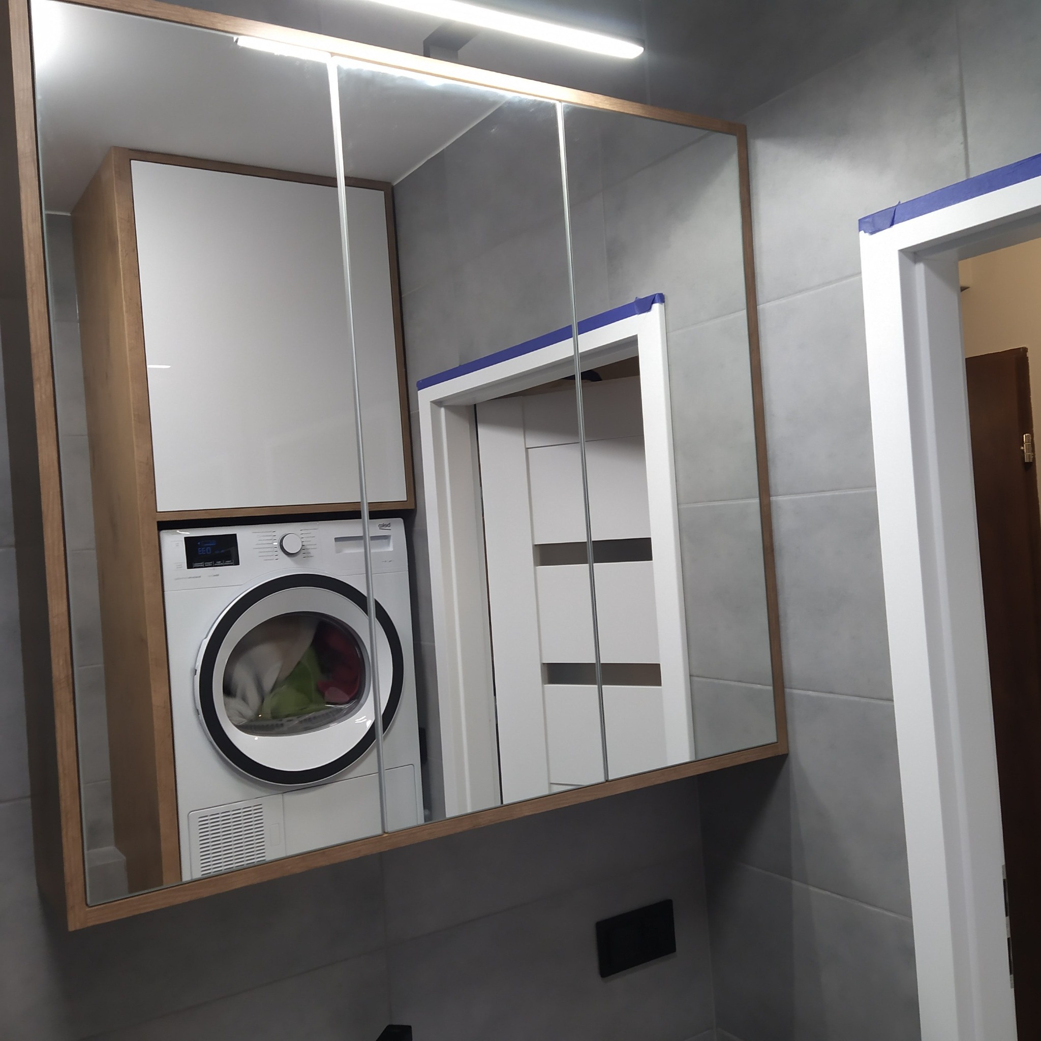 Łazienka z szarą płytką, zabudowana pralka w szafce z lustrzanymi drzwiczkami, widoczna część futryny drzwi z niebieską taśmą malarską.