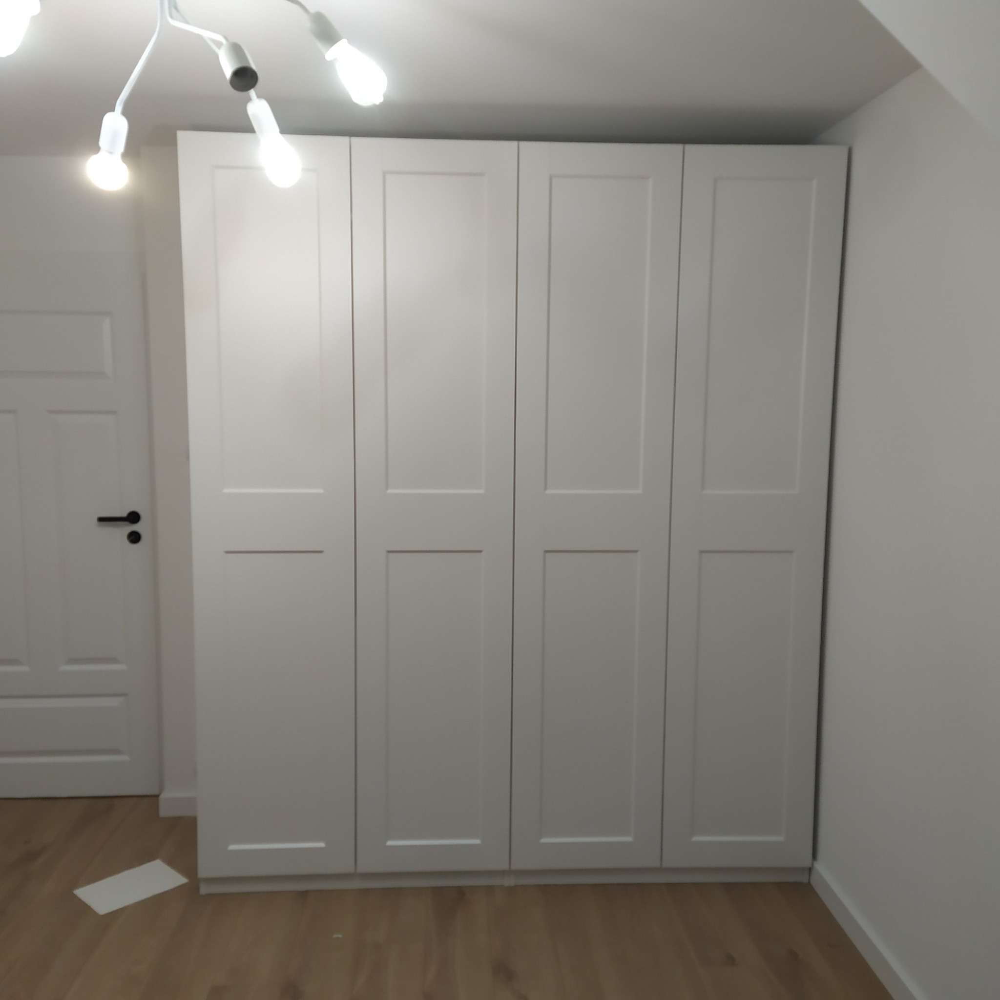 Zamontowana, biała szafa czterodrzwiowa z panelowymi drzwiami w pokoju ze skośnym sufitem, widoczna obok biała ściana i drzwi z czarną klamką, na podłodze leży biała kartka.