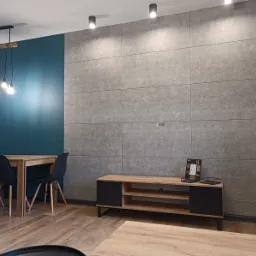 Nowoczesne wnętrze: ściana wyłożona szarymi płytami imitującymi beton, drewniany stół z czarnymi krzesłami, szafka RTV z elementami drewna i czerni, oraz minimalistyczna lampa wisząca z trzema...