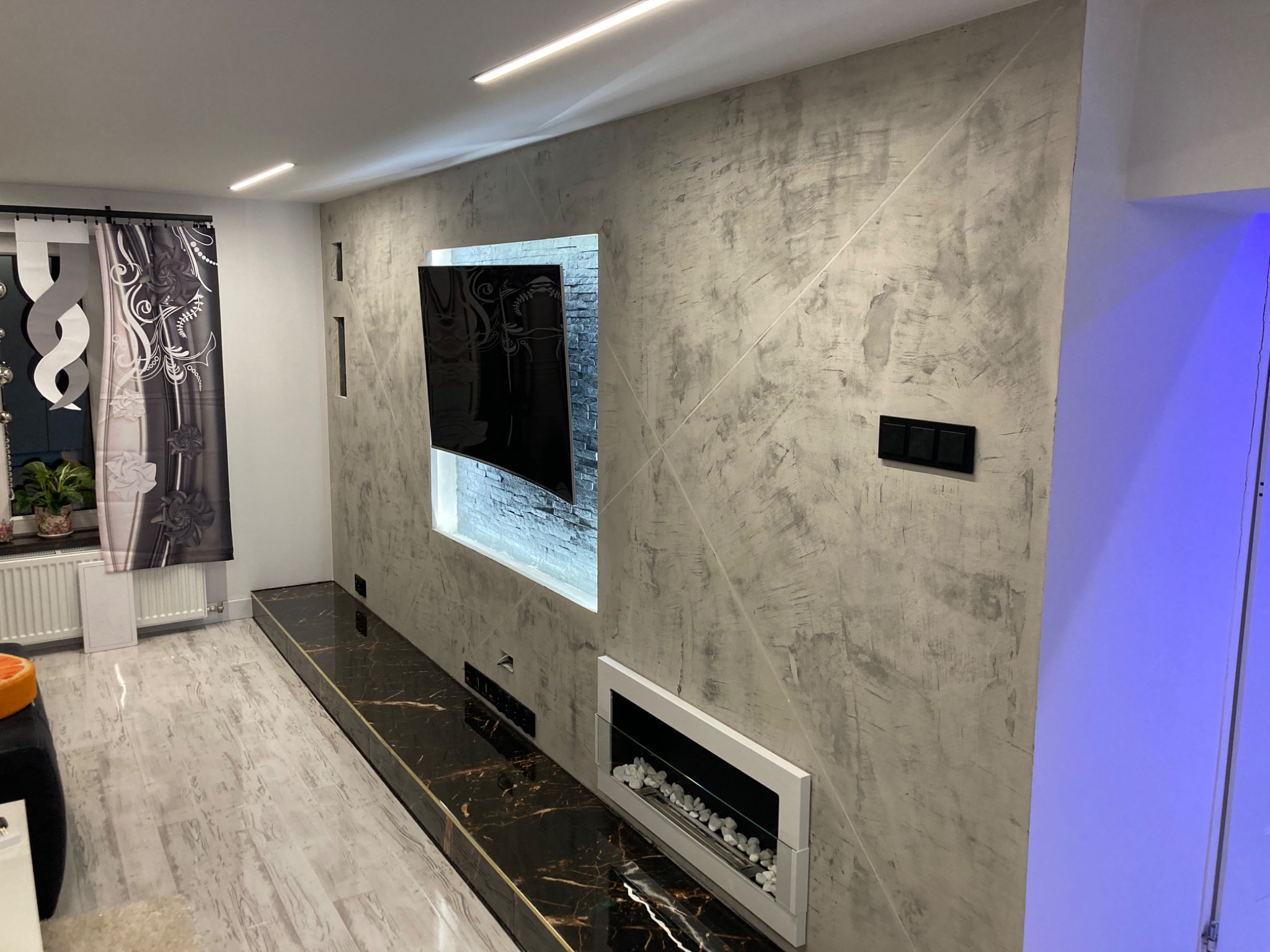 Ściana w salonie z dekoracyjną szarą strukturą, podświetlonym wnęką z telewizorem, imitacją kominka z białym obramowaniem i czarnym blatem z marmurowym wzorem.