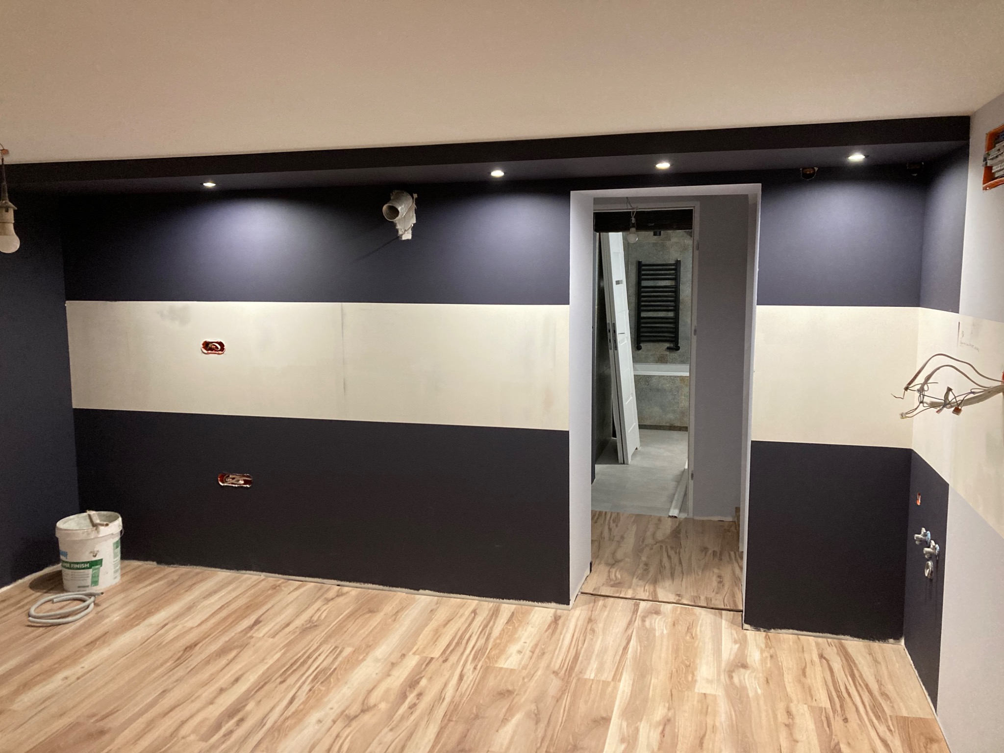 Wnętrze w trakcie malowania, z ciemnografitową dolną i górną częścią ścian oraz jasną środkową, widoczne puszki z farbą, instalacje elektryczne i fragment łazienki w tle.