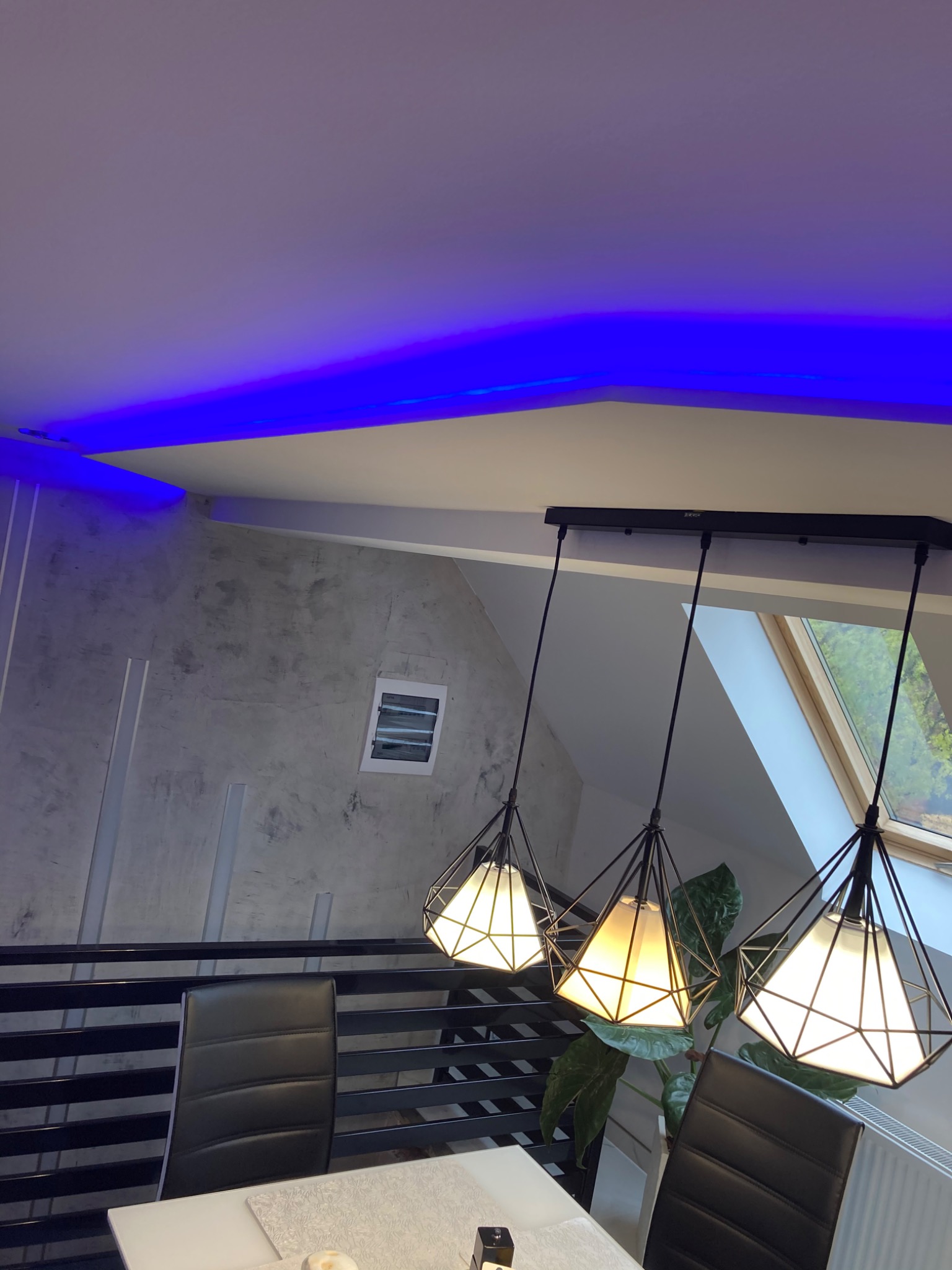 Nowoczesna jadalnia z trzema wiszącymi lampami w kształcie diamentów, oświetlenie LED w kolorze niebieskim na suficie, ściana z efektem betonu i czarne krzesła przy białym stole.